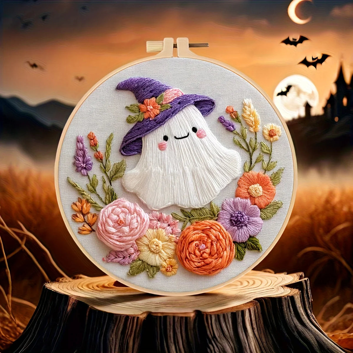 A circular embroidery hoop displays a whimsical embroidered design of a smiling white ghost wearing a purple witch hat adorned with flowers, surrounded by colorful embroidered roses and foliage.