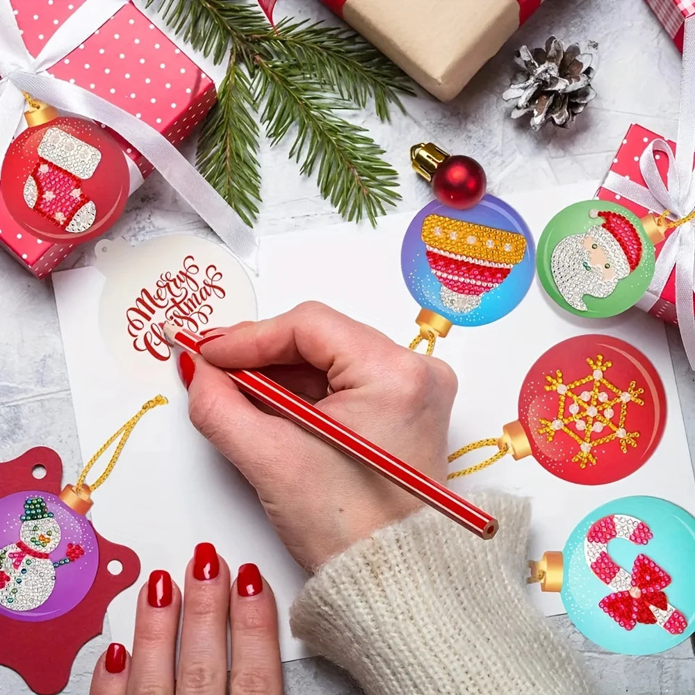 A hand with red nail polish writes on a white Christmas ornament tag with a red pen, surrounded by festive decorations including colorful hanging ornaments, wrapped gifts, pine branches, and a pinecone