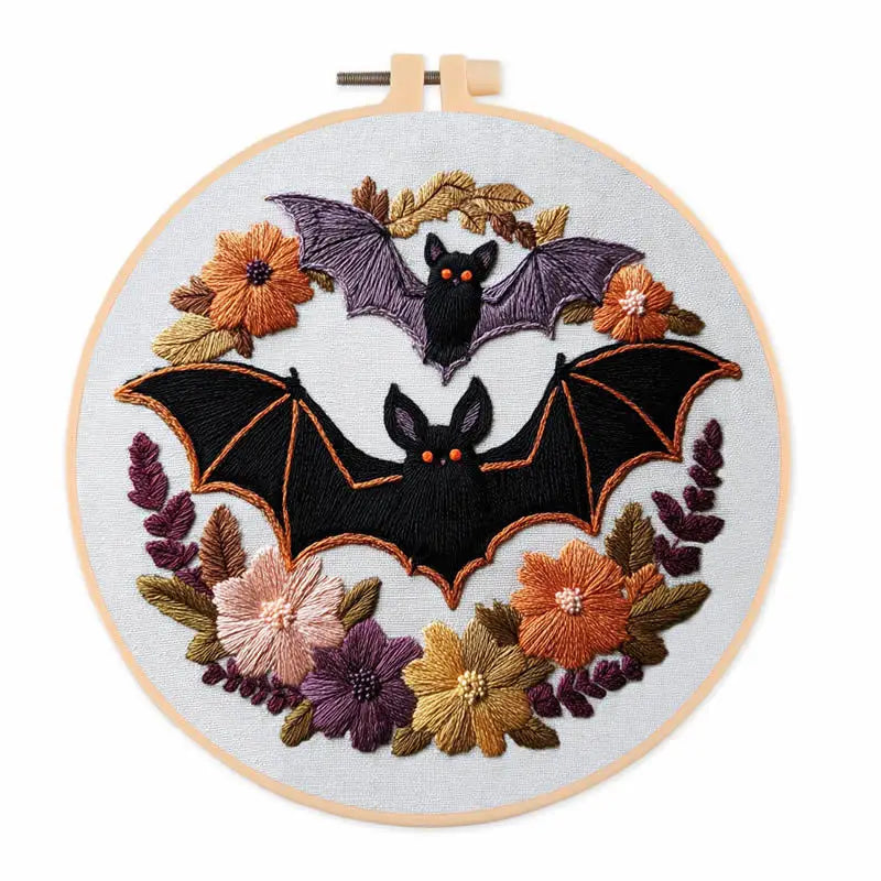 A circular embroidery hoop containing a completed embroidered design of two black bats surrounded by orange, purple, and gold flowers on a white fabric background.