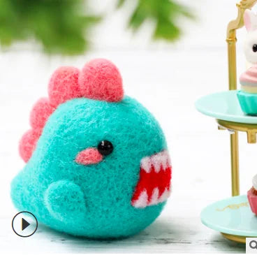 A turquoise needle felted monster with a red spikes going down it's back, and it's mouth open red and white sharp monster teeth