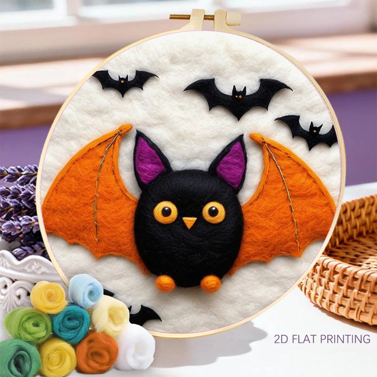 A circular embroidery hoop displays a cute bat-themed needlepoint or felt artwork. The design features a black bat face with yellow eyes and a small orange nose, purple inner ears, and large orange wings, set against a white background with three small black bats.