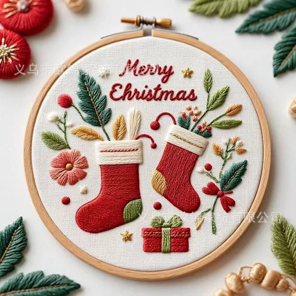 A round wooden embroidery hoop containing a festive Christmas stitched design with two red stockings, green leaves, red berries, a small gift box, and the words “Merry Christmas” in red script. The hoop is centered on a light background with decorative green leaves and red ornaments around it.
