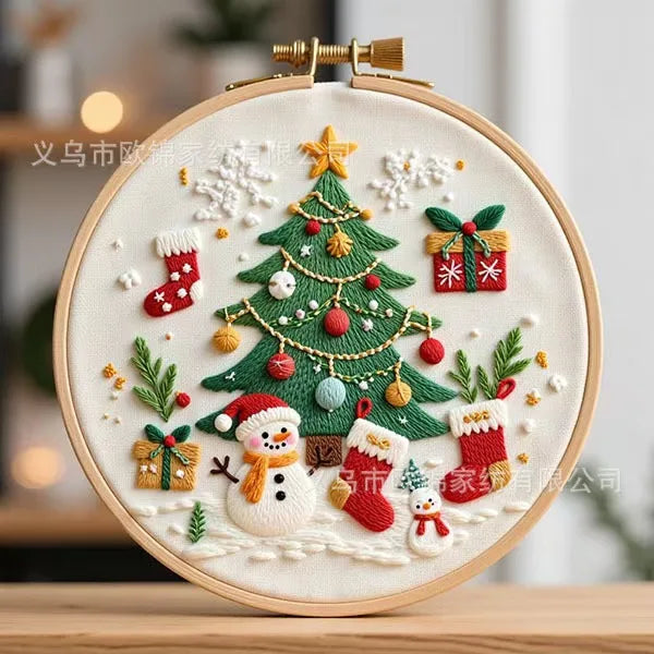 A round embroidery hoop containing a festive Christmas-themed embroidered design with a green tree, snowman, Santa, stockings, gifts, and snowflakes on a cream fabric background.