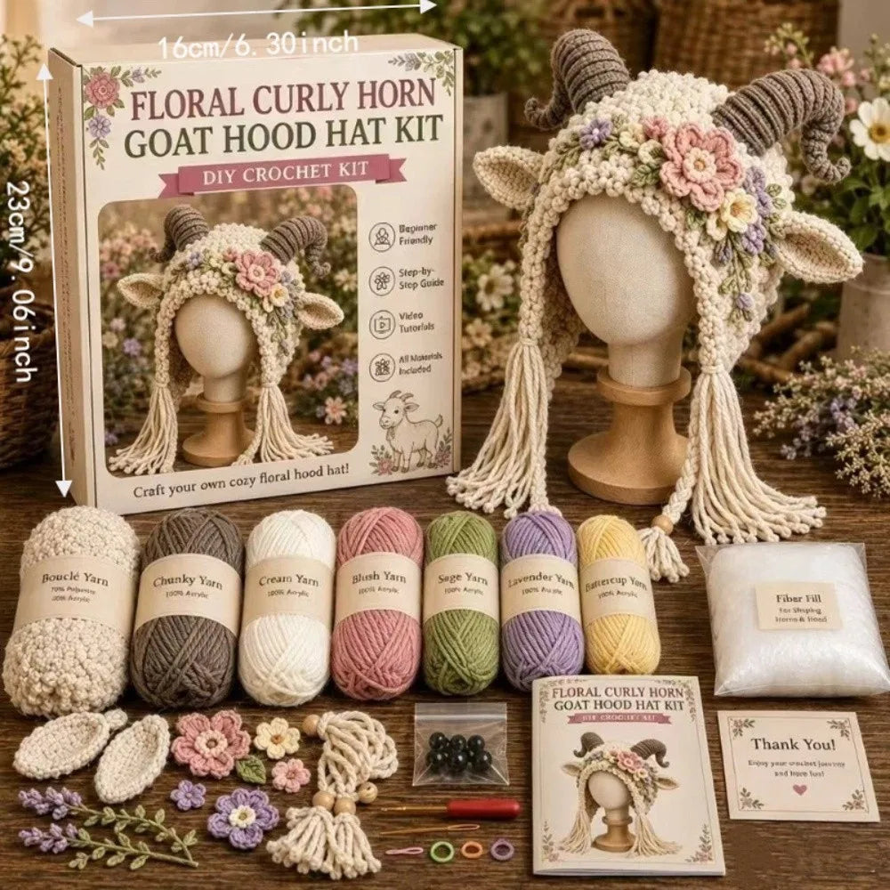 Floral curly horn goat hood hat kit shown with its retail box, yarn balls, crochet tools, flowers, and a finished crocheted hat displayed on a mannequin head. The scene is styled on a wooden surface with floral decor in the background.