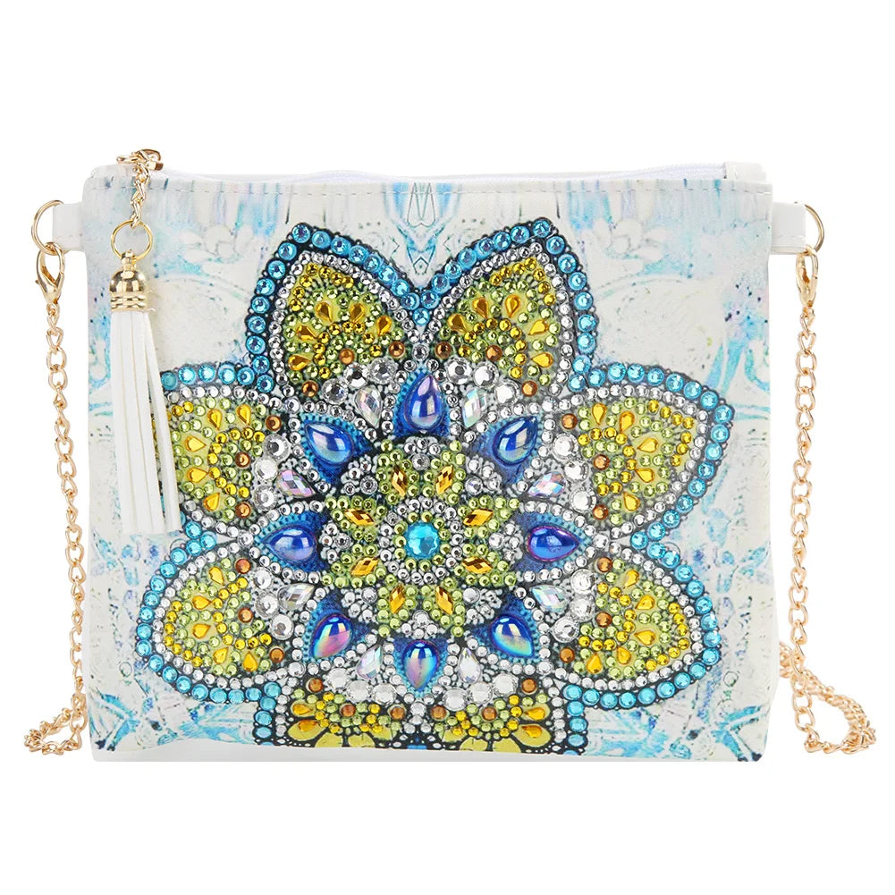 A rectangular crossbody clutch bag with a white and light blue marbled background, featuring a large central mandala design embellished with yellow, blue, and silver rhinestones. It has a white tassel charm and a gold-tone chain strap.