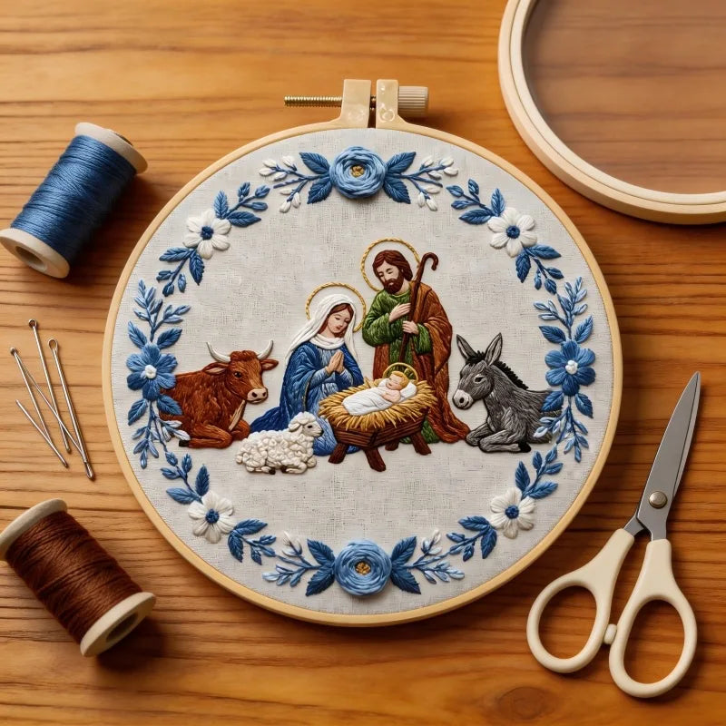 A circular embroidery hoop, featuring a nativity scene stitched with blue, white, green, brown, and gold thread. 