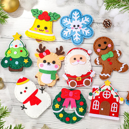 A flat lay of colorful felt Christmas-themed cutouts on a whitewashed wood surface, including a yellow bell, blue snowflake, green Christmas tree, reindeer, Santa Claus, gingerbread man, snowman, wreath, and red house, with small pinecones and evergreen branches around them.