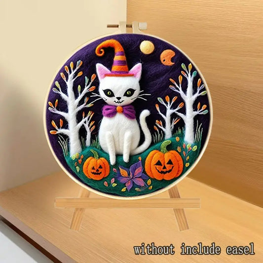 A circular needle felted art piece of a white cat in a witch hat surrounded by pumpkins, trees, and a purple night sky