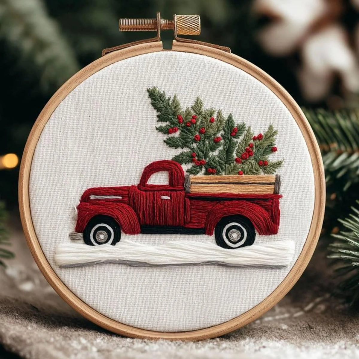 A circular embroidery hoop displays a festive stitched design of a red pickup truck carrying a green Christmas tree with red berries, set against a snowy white ground. 