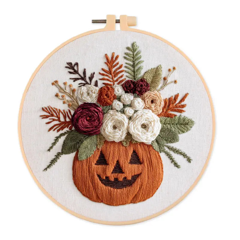 A circular embroidery hoop displaying a floral pumpkin design. The orange pumpkin features an embroidered  jack-o'-lantern face and is filled with a bouquet of flowers in white, burgundy, orange, and green, all set against a plain white background.