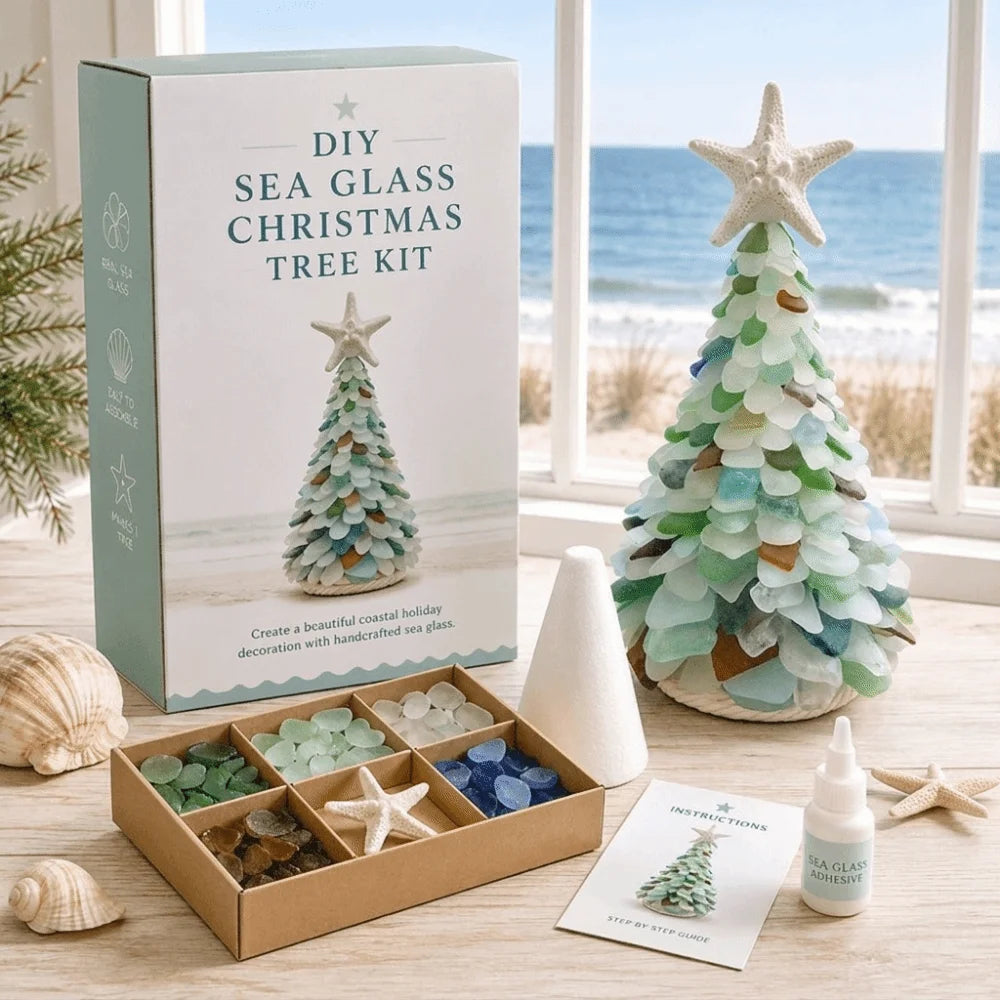 DIY Sea Glass Christmas Tree Kit displayed on a wooden table by a window with a beach view. The kit includes a mint green and white instruction box, a completed sea glass tree decorated with green, blue, and beige stones, a cardboard tray of sea glass beads, a white cone, a small bottle of glue, and an instruction card.