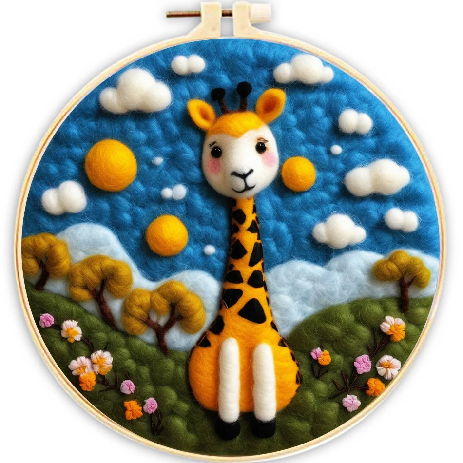 Needle felted giraffe sitting on a green hillside with flowers, a blue sky with white clouds in a hoop frame