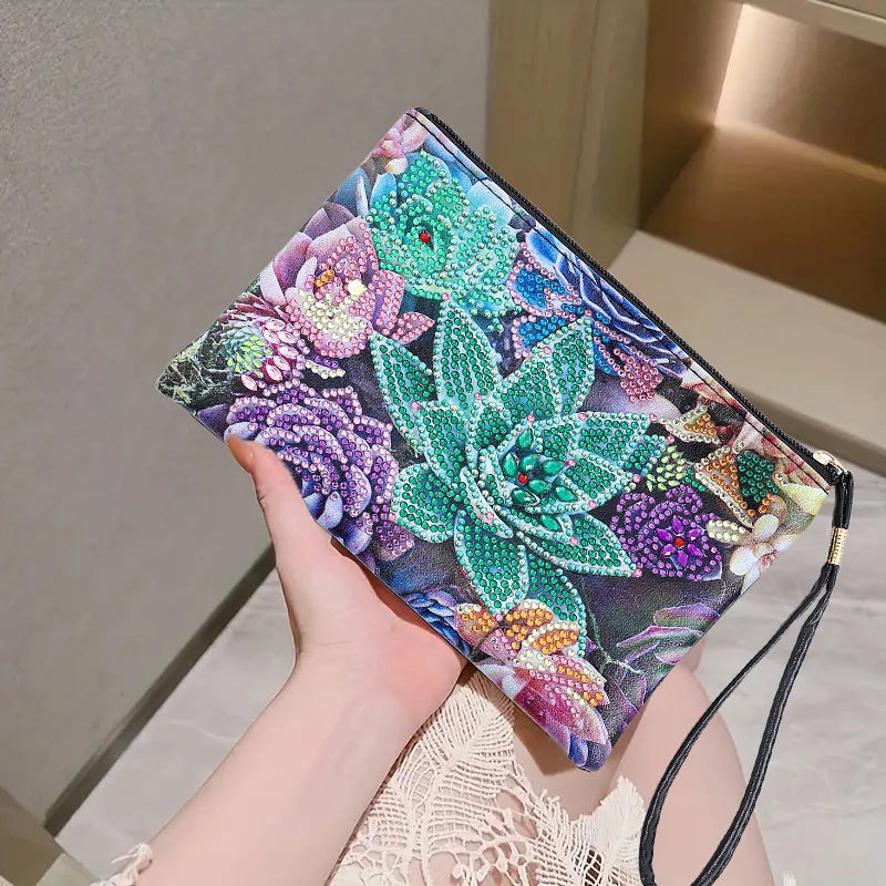 A hand holds a zippered clutch bag decorated with a colorful diamond art lotus patterns. The clutch features a black wrist strap 