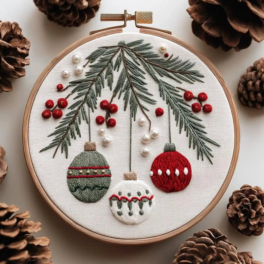 A circular embroidery hoop displays a festive Christmas-themed embroidery design featuring pine branches, red berries, and three hanging ornaments. The hoop is styled with pinecones on a light beige background.