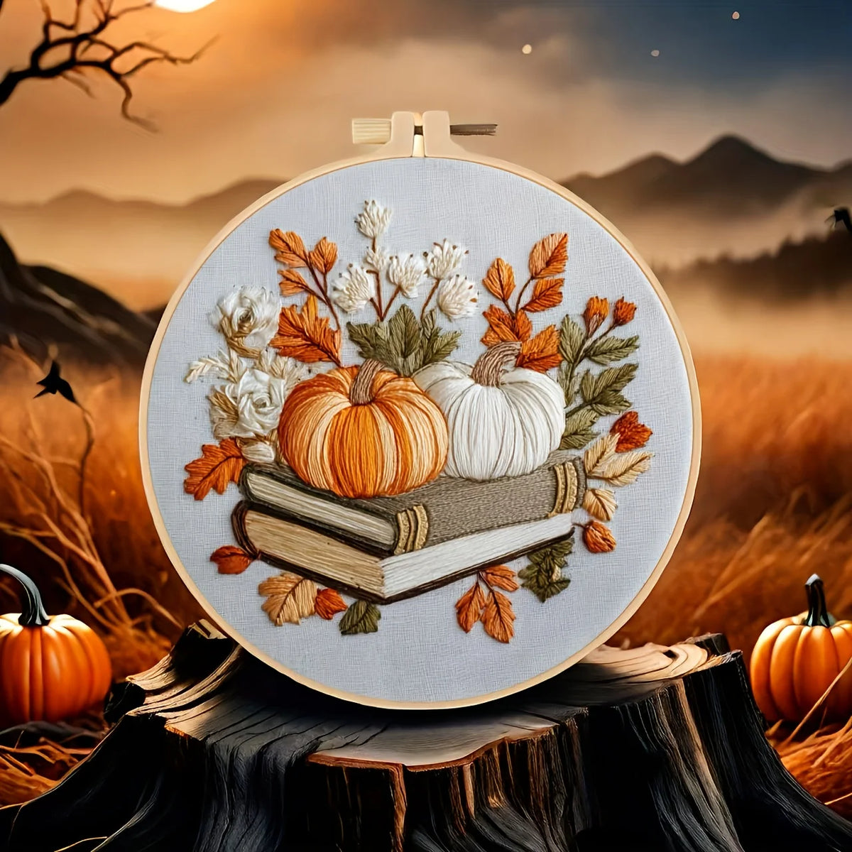 A circular embroidery hoop displays a detailed autumn-themed embroidered design featuring two pumpkins, white flowers, and stacked books. 