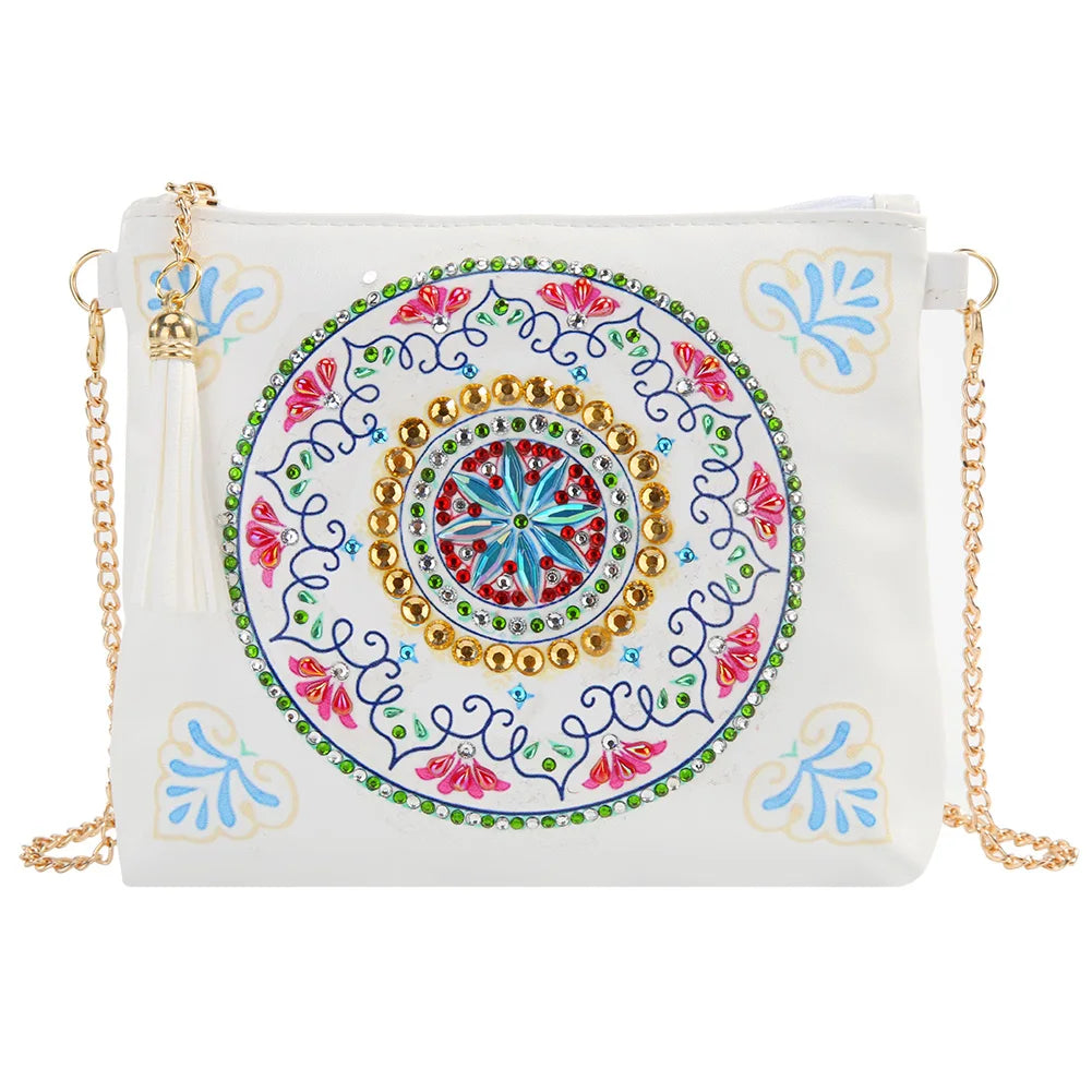 A white faux-leather crossbody bag featuring a large, colorful circular mandala design on the front, embellished with gold beads, green rhinestones, and pink floral details. It has a white tassel charm and a gold-tone chain strap
