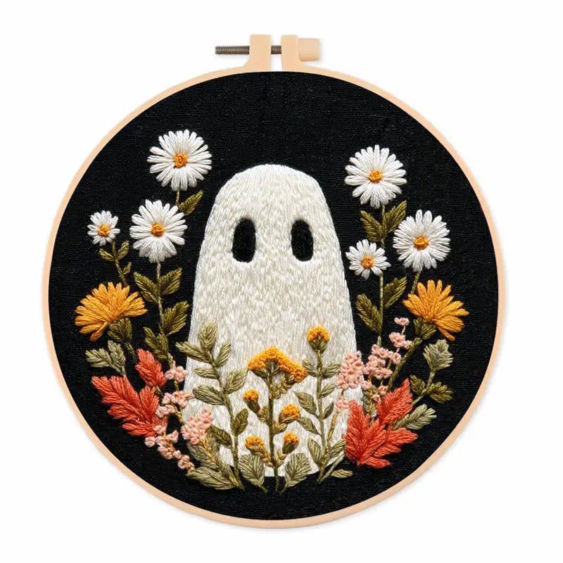 A circular embroidery hoop containing a black fabric background with a white ghost motif surrounded by embroidered white daisies, yellow flowers, green leaves, and orange-red leaves.