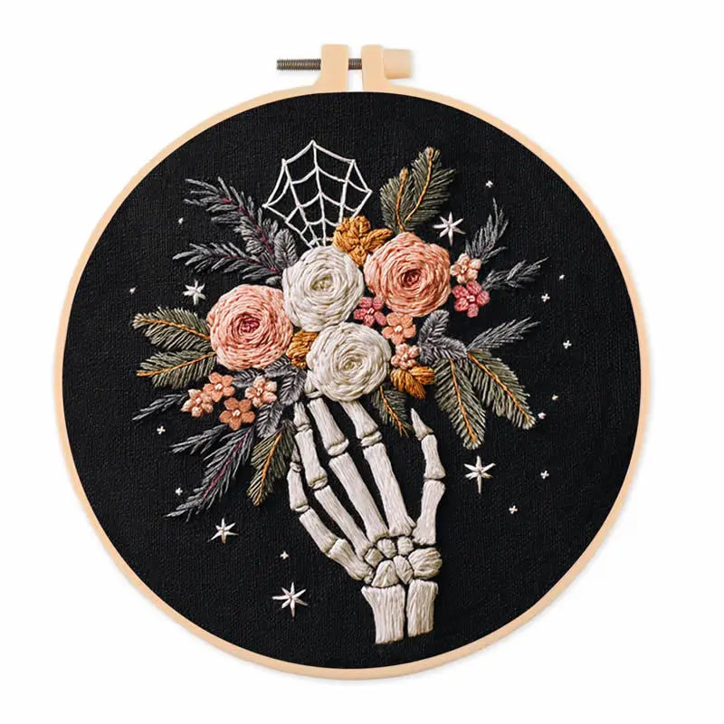 A circular embroidery hoop containing a black fabric backdrop with a hand-embroidered design of a skeleton hand holding a bouquet of pink, white, and orange flowers with green leaves, a spiderweb motif, and small white stars.