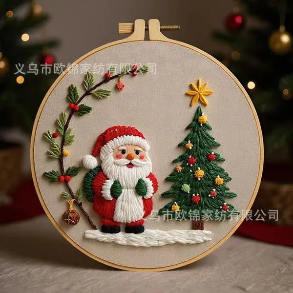 A circular embroidery hoop featuring a festive Christmas design with a stitched Santa Claus, decorated Christmas tree, holly branch, and gold star, shown against a beige background.