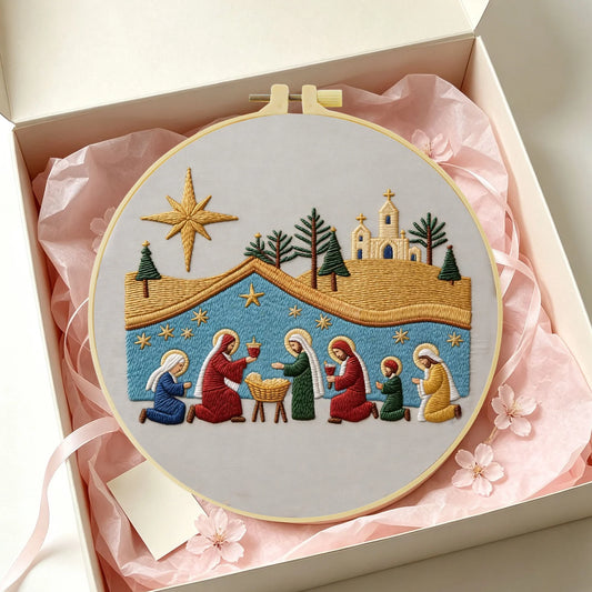 A circular embroidery hoop containing a colorful Christmas nativity scene stitched design, presented inside an open cream gift box with pink tissue paper and ribbon.