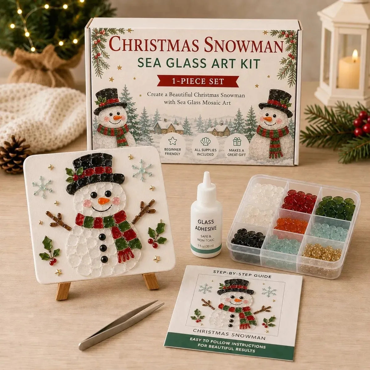 Christmas Snowman Sea Glass Art Kit displayed on a tabletop with its box, finished mosaic tile on a small easel, glass adhesive bottle, tray of colorful sea glass beads, tweezers, and a step-by-step guide. Festive holiday decor including pine branches, a candle, and a knitted blanket is visible in the background.