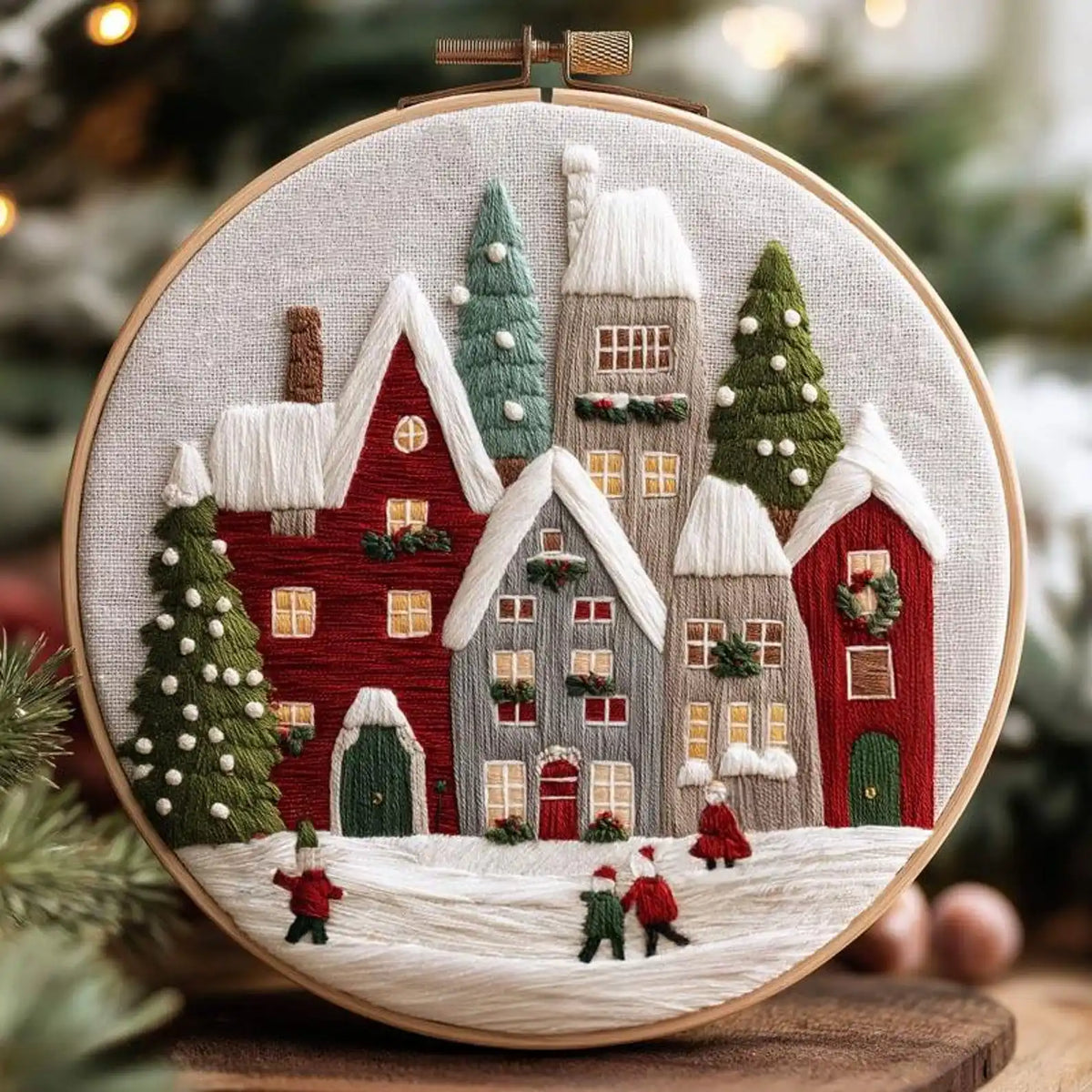 A circular embroidery piece in a wooden hoop showing a cozy winter village scene with snow-covered houses, evergreen trees, and small figures in the foreground, photographed against a softly blurred holiday background.
