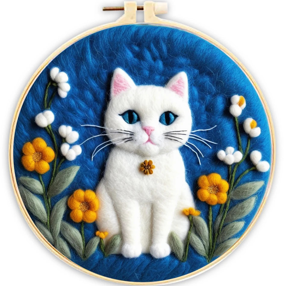 A circular needle felted art piece featuring a white cat with blue eyes and a small yellow flower collar, surrounded by yellow and white flowers on a textured blue background, framed in a wooden hoop.